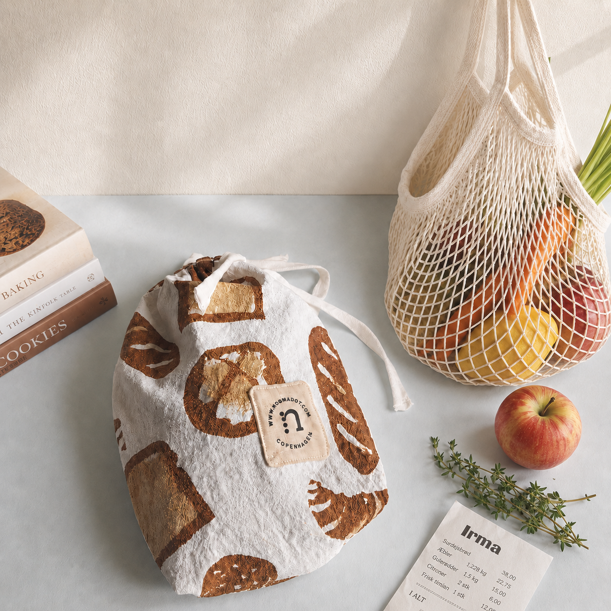 Printed cotton bakery bag styled with a reusable mesh bag of fruit, apple, herbs, receipt and baking book on a light neutral background.