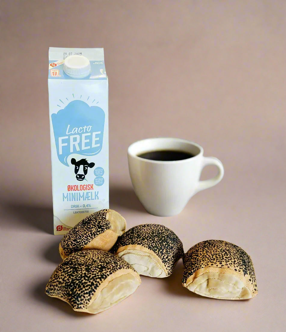 Carton of lactose-free organic milk and a white coffee cup positioned behind four toy Copenhagen poppy-seed buns on a soft beige background.
Karton med laktosefri økologisk minimælk og hvid kaffekop placeret bag fire legeboller med birkes på neutral beige baggrund.