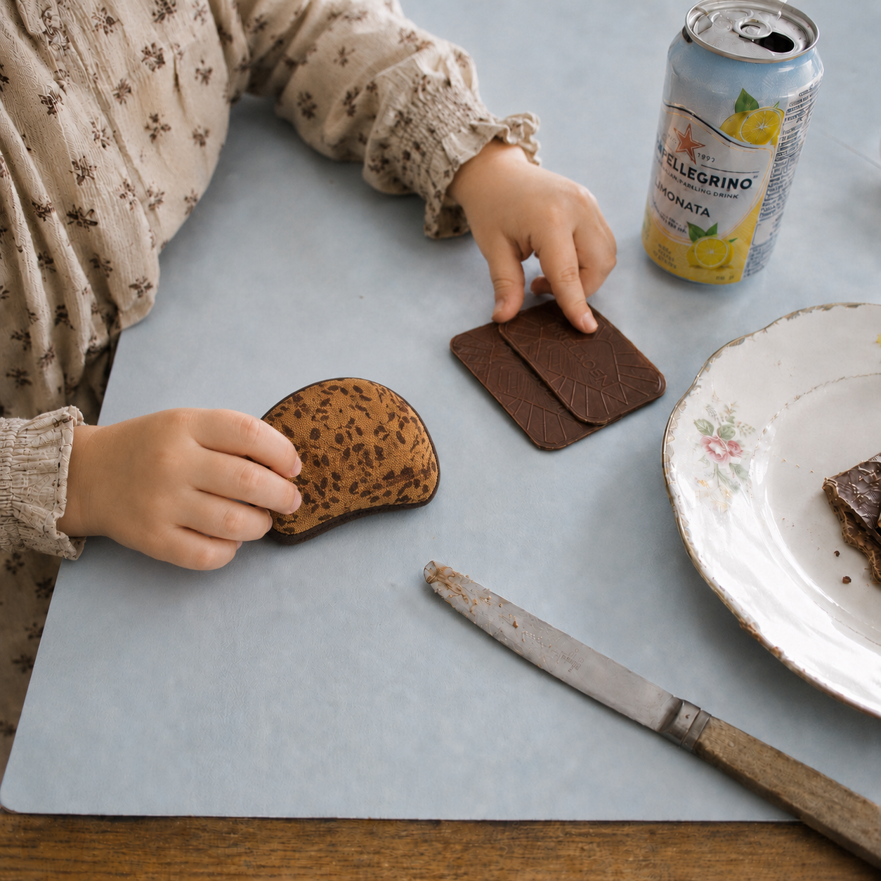 Child playing with a handmade fabric rye bread slice at a blue table with chocolate biscuits, vintage plate, knife and lemonade can.
