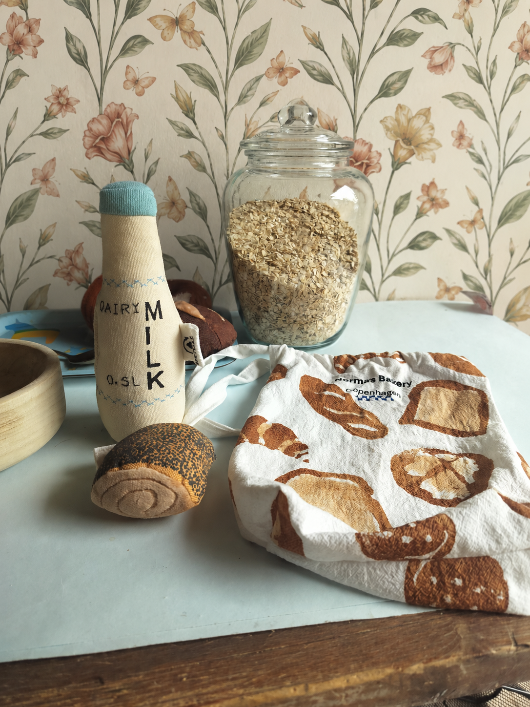 Play Food Milk bottle, jar of oats, and bread-themed towel on a table with floral wallpaper.