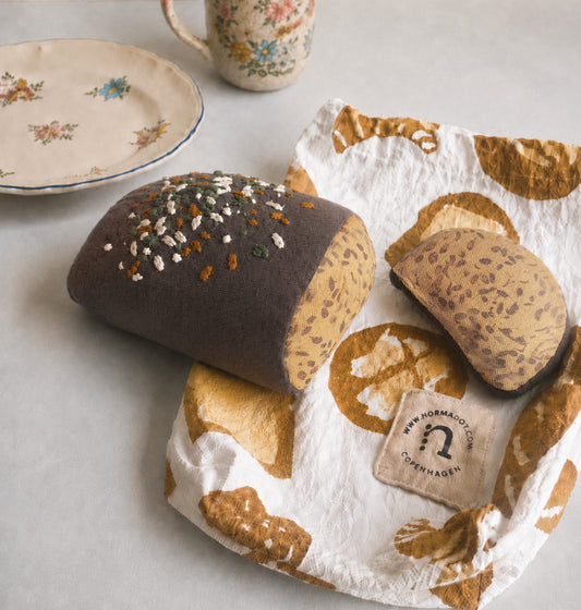Handmade fabric rye bread loaf with one slice on a printed cotton bakery bag, styled with vintage plate and cup on a light neutral background.