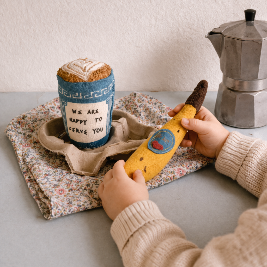 Person holding a banana with a sticker, next to a coffee cup and Moka pot on a table.