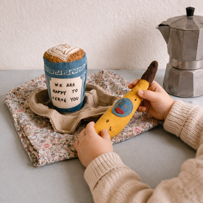 Person holding a banana with a sticker, next to a coffee cup and Moka pot on a table.