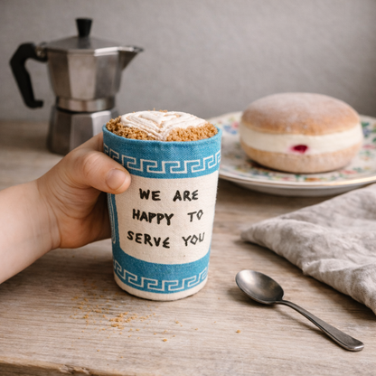 Hand holding a coffee cup fabric toy with a decorative sleeve, surrounded by a donut and a coffee maker on a wooden table.