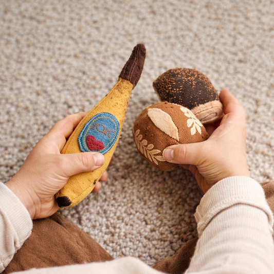 Two hands holding a yellow banana-shaped toy and a brown bakery rollshaped toy on a textured surface.