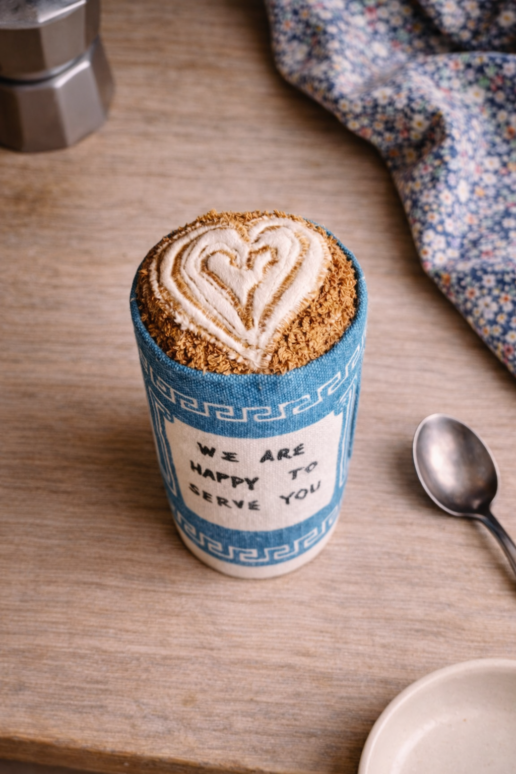 Coffee cup Fabric Toy with a heart-shaped latte art on a wooden table with a spoon and floral cloth.