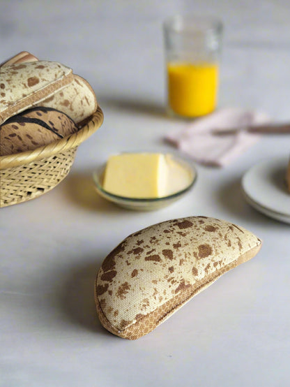 "A realistic fabric sourdough bread toy placed on a light surface, with a basket of similar toy breads, a glass of orange juice, and a dish of butter in the background."
"Et realistisk stykke legetøjs-surdejsbrød i stof ligger på en lys overflade, med en kurv med lignende legetøjsbrød, et glas appelsinjuice og en tallerken med smør i baggrunden."