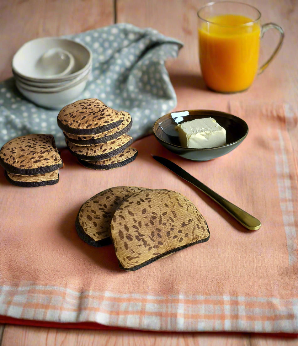 Fabric toy rye bread slices on a pink cloth with a butter dish, knife, stacked bowls, and a glass of orange juice – pretend play breakfast scene.
Stoflegebrødskiver af rugbrød på en lyserød dug med smørskål, kniv, stablede skåle og et glas appelsinjuice – fantasifuld morgenmadsscene.