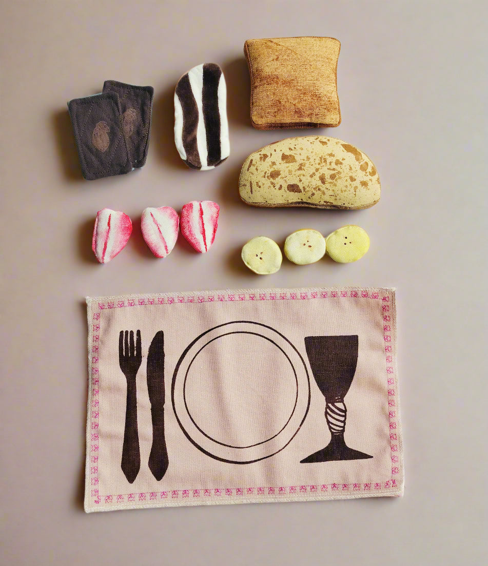 Flat lay of a handmade fabric play food set including sandwich chocolate slices, chocolate spread toast, cinnamon toast, sourdough bread, strawberry slices, banana slices, and an embroidered placemat with a printed table setting.
Fladtlæggende visning af håndlavet legemad i stof med pålægschokolade, chokoladepålæg på toast, kaneltoast, surdejsbrød, jordbærskiver, bananskiver og en broderet dækkeserviet med trykt opdækning.