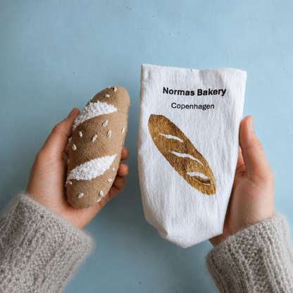 
Hands holding a handmade fabric baguette and printed cotton bakery bag against a light blue background.
