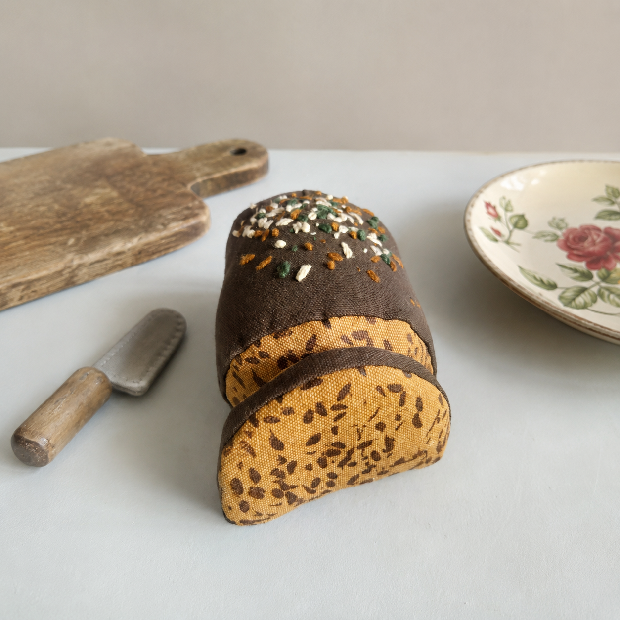 
Handmade fabric rye bread loaf with one slice, styled with a small wooden butter knife, cutting board and vintage floral plate on a light neutral background.

