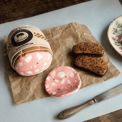 Handmade fabric mortadella, mortadella slice and rye bread slices styled on brown crinkled paper with a vintage knife and floral plate.