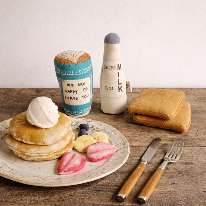 Stack of play food pancakes with syrup, fruit, and whipped cream on a plate with a milk bottle and coffee cup in the background.