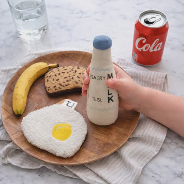 Child playing with handmade breakfast play food, including a fabric milk bottle, banana, fried egg and bread on a wooden tray.