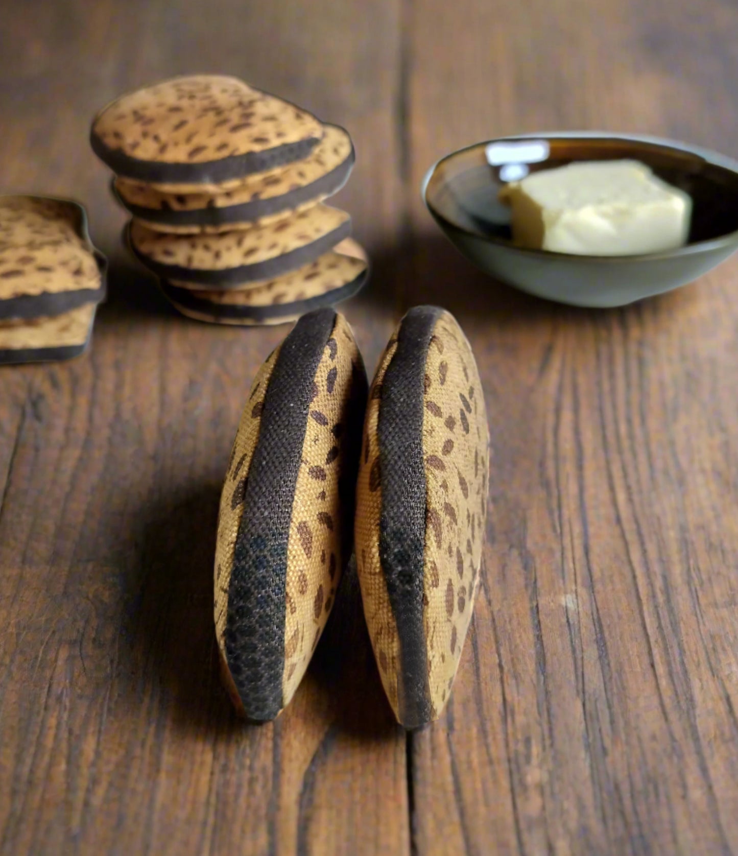 Two upright fabric rye bread toy slices on a wooden surface with stacked slices and a bowl of butter in the background.
To stående stoflegebrødskiver af rugbrød på en træoverflade med stablede skiver og en skål med smør i baggrunden.