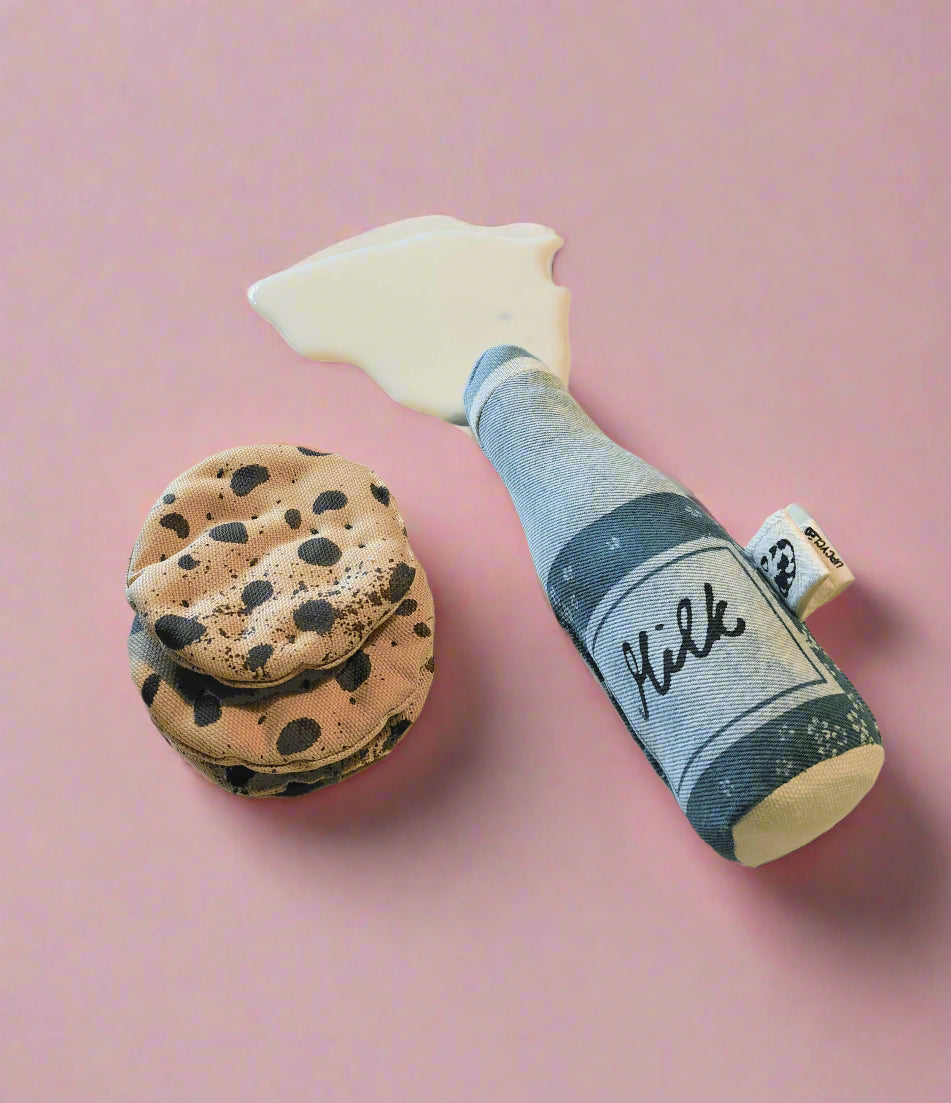 “Upcyklet denim-mælkeflaske med spilledråbe af ‘mælk’ ved siden af tre stofcookies på lyserød baggrund.”
“Upcycled denim milk-bottle soft toy with a splash of ‘milk’ beside three fabric cookies on a pink background.”